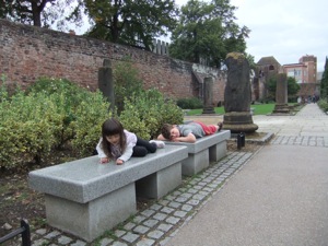 チェスター 真由喜とお友達がローマ時代の柱のまえに新しい意志のベンチにうつぶせになる。背景二は、チェスターの中世の塀が見える。