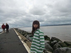 ウィラル半島の海岸 大きなセーターを着た真由喜が灰色の空の下で茶色の海の前に立つ