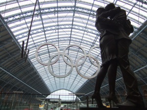 銅像の向こうに五輪のマークとホームが見える