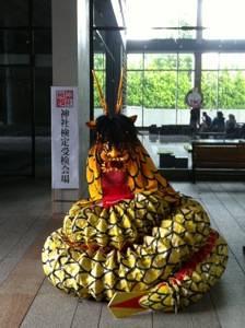 大学の建物の前に八岐大蛇の衣装が建っている。背景には検定会場の看板がある