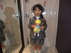 幼稚園の制服を着て、幼稚園児の姿の人形を持っている真由喜